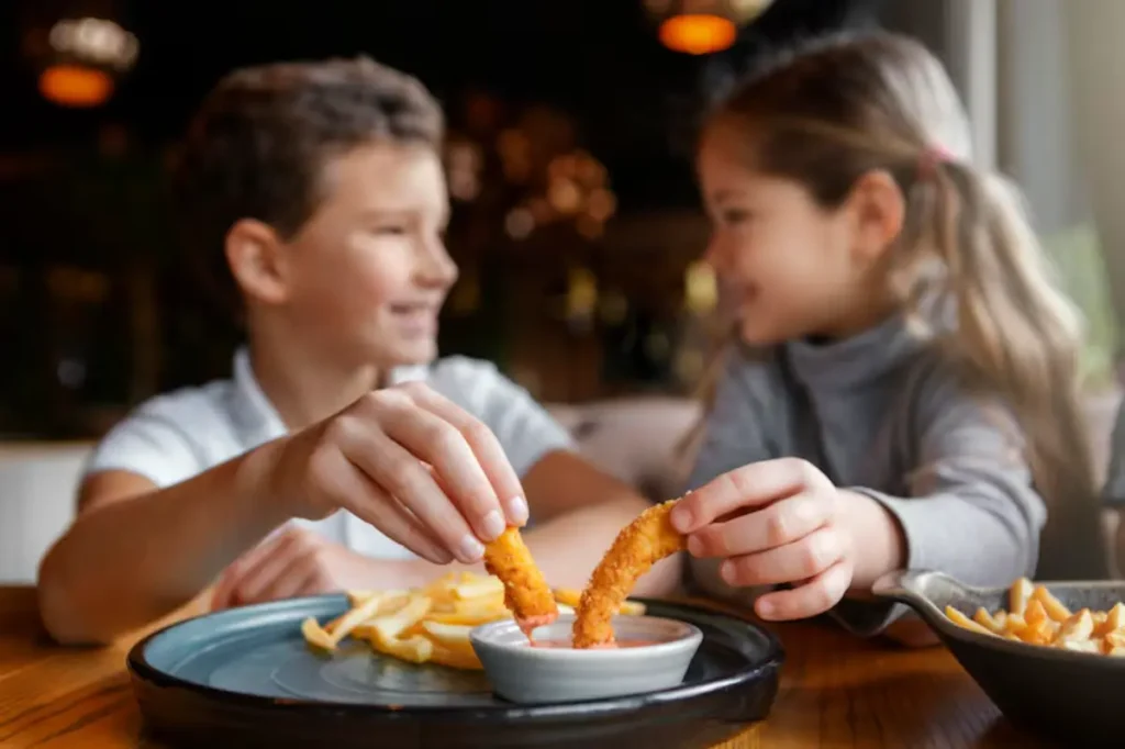 Two children are enjoying a meal, dipping crispy shrimp into a small dish of sauce, with fries on a plate nearby.