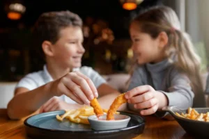 Two children are enjoying a meal, dipping crispy shrimp into a small dish of sauce, with fries on a plate nearby.