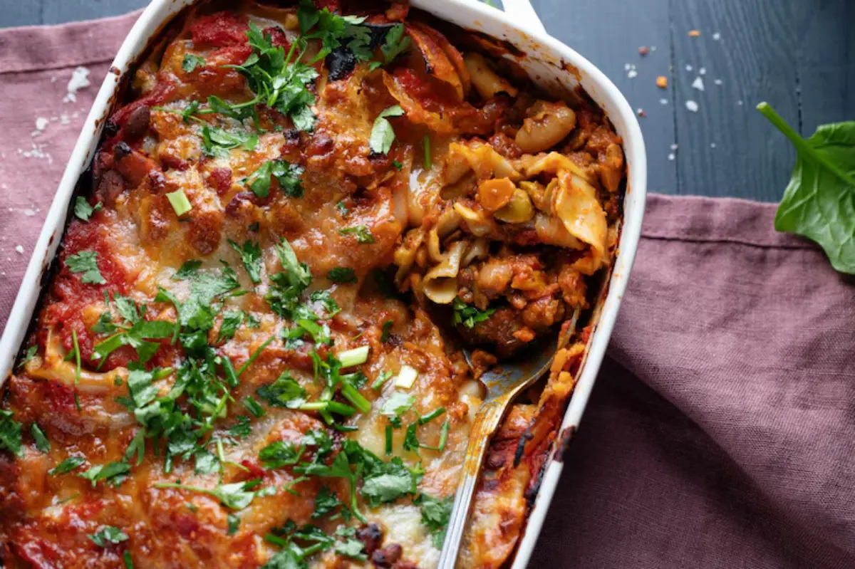 A cheesy baked pasta dish topped with fresh herbs, partially dished out and served in a white casserole dish on a purple cloth.