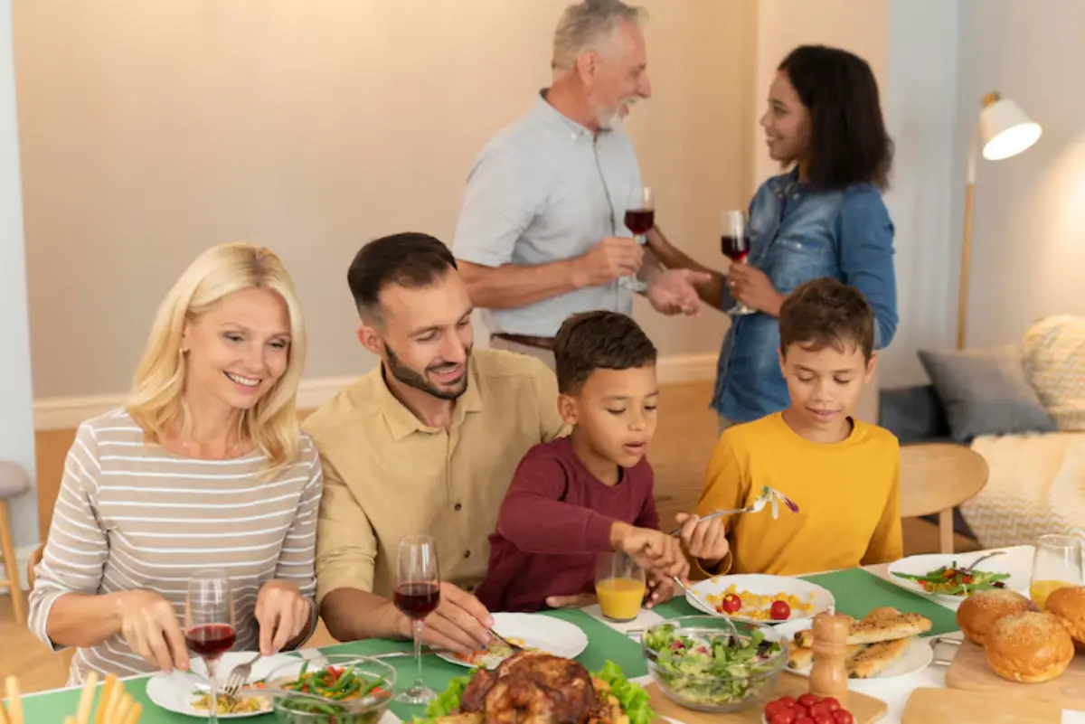 A family gathers around a dining table, enjoying a meal with various dishes like salad and roasted chicken, while others converse nearby.