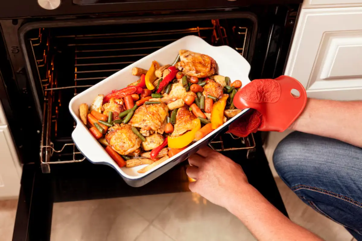 A person wearing a red oven mitt pulls a baked dish of chicken and colorful vegetables from the oven.