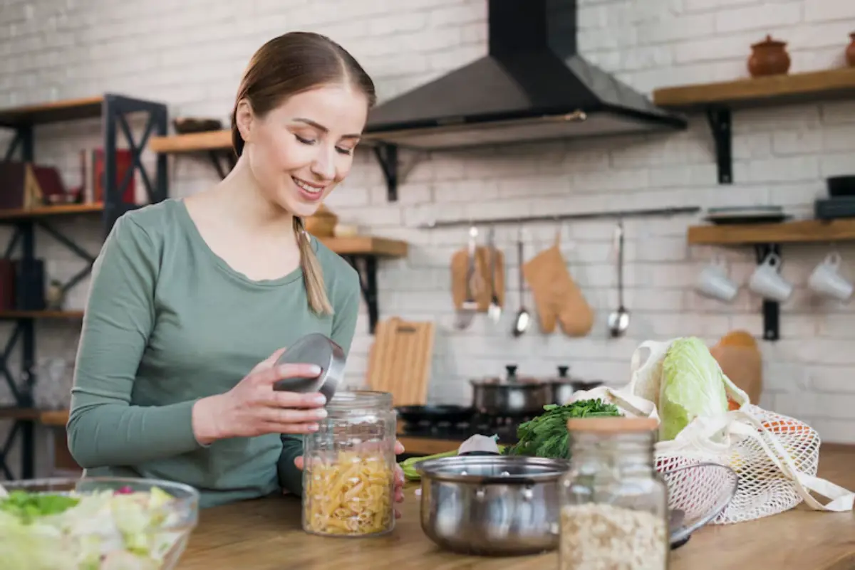 A woman in a green shirt organizes pasta in a jar while surrounded by fresh vegetables and kitchen tools in a bright kitchen.