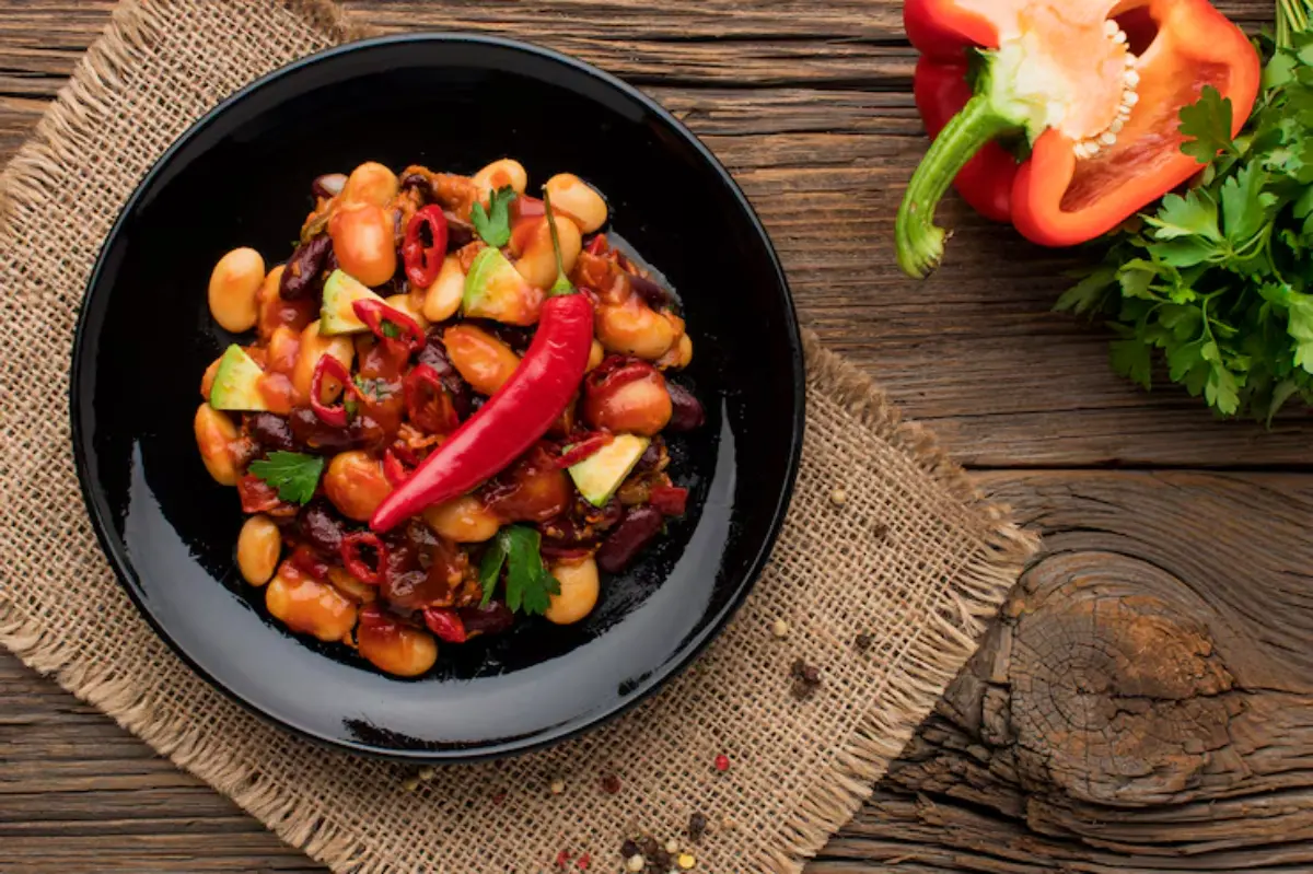 A vibrant dish of mixed beans and vegetables garnished with red chilies, served on a black plate over a rustic wooden table.