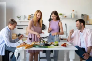A group of friends enjoys a meal together at a bright kitchen table, featuring various dishes, wine, and fresh ingredients.