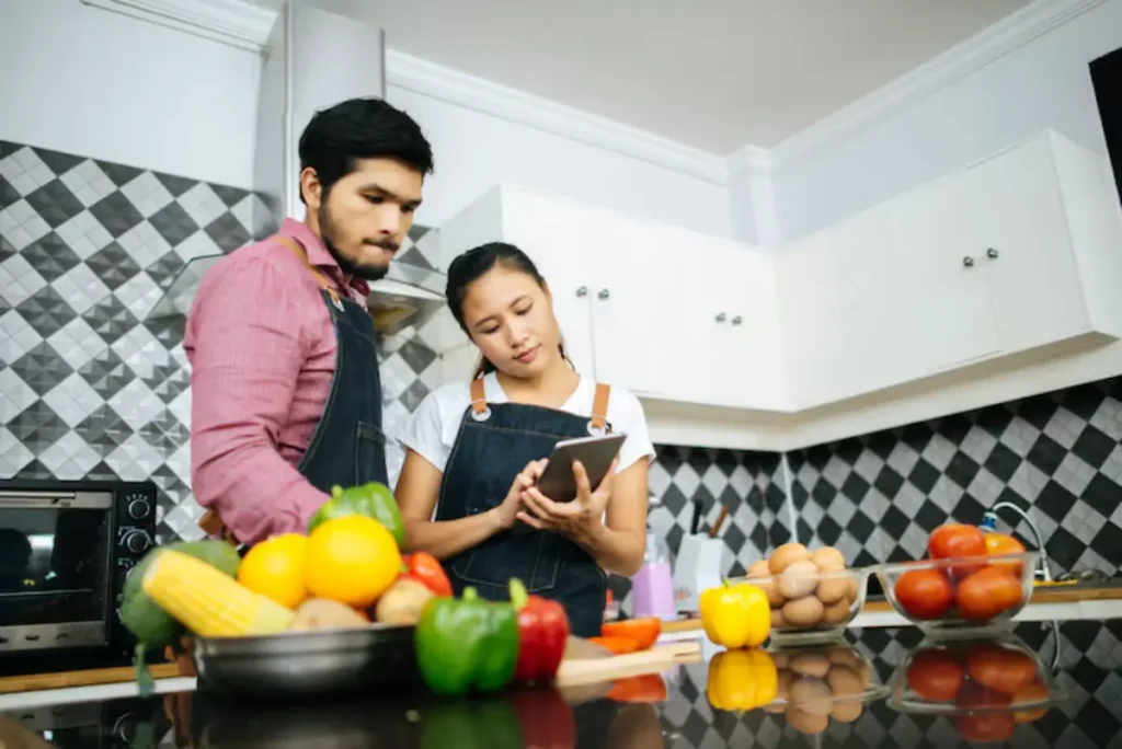 A couple in matching denim aprons stands in a modern kitchen, discussing recipes on a tablet, surrounded by fresh vegetables and eggs.