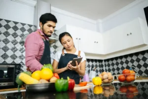 A couple in matching denim aprons stands in a modern kitchen, discussing recipes on a tablet, surrounded by fresh vegetables and eggs.