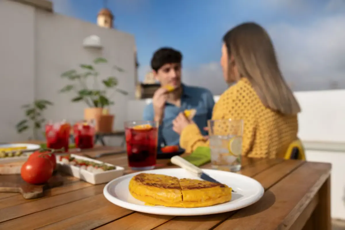 A plate of golden tortilla with a knife on a wooden table, surrounded by drinks and fresh ingredients, set on a sunny rooftop.