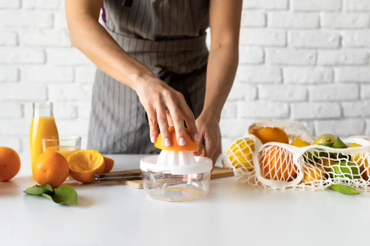 A person squeezes an orange using a manual juicer, with fresh oranges, juice bottles, and a vibrant kitchen backdrop.