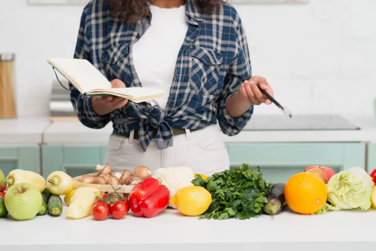 A person in a flannel shirt holds a notebook and pen, surrounded by fresh vegetables and fruits on a kitchen counter.