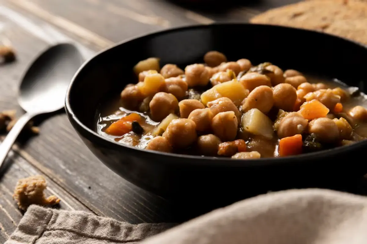 A black bowl filled with hearty chickpea stew, featuring diced vegetables on a wooden table, accompanied by a spoon and bread.