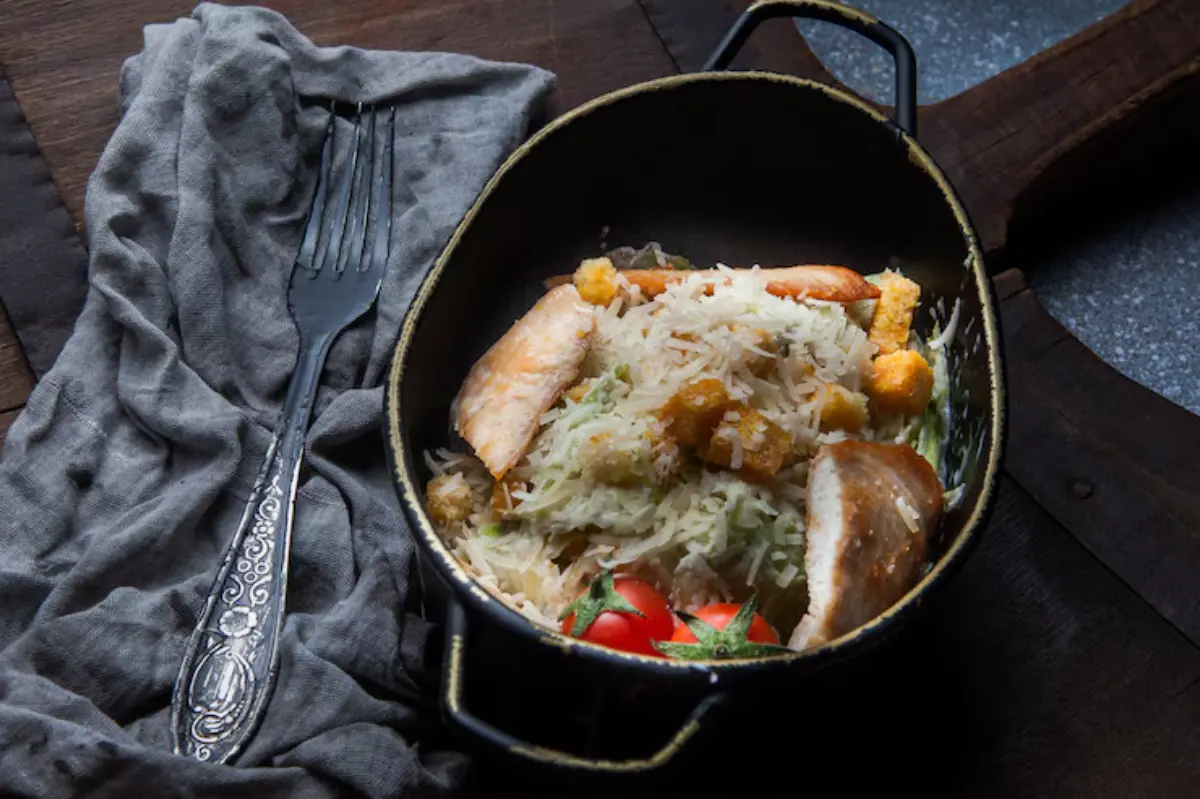 A bowl of fluffy rice garnished with vegetables, fried chicken pieces, and cherry tomatoes, accompanied by a silver fork on a gray cloth.