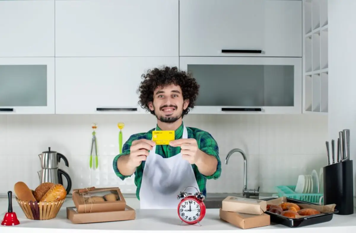 A person in a kitchen holding a yellow card, surrounded by baked goods, a red alarm clock, and kitchen utensils on a countertop.