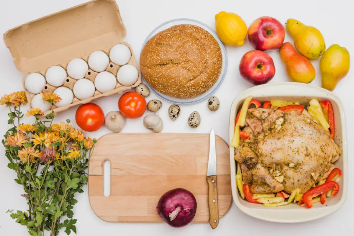 A layout of fresh ingredients including a whole chicken, eggs, vegetables, fruits, a loaf of bread, and flowers on a wooden cutting board.