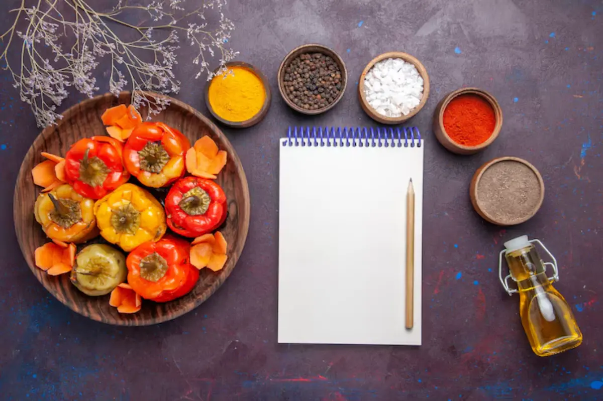 A wooden bowl filled with colorful peppers beside spices, a notepad, and a bottle of oil on a dark textured surface.