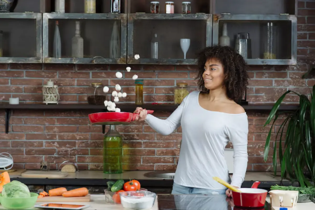 A woman in a white shirt tosses mushrooms from a red pan in a rustic kitchen filled with fresh vegetables and glass jars.