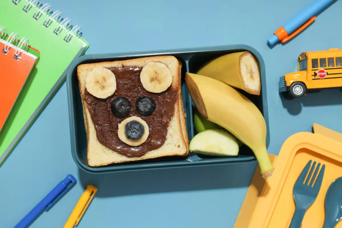 A playful lunchbox featuring a sandwich decorated as a bear's face with banana and blueberry, surrounded by fruits and school supplies.
