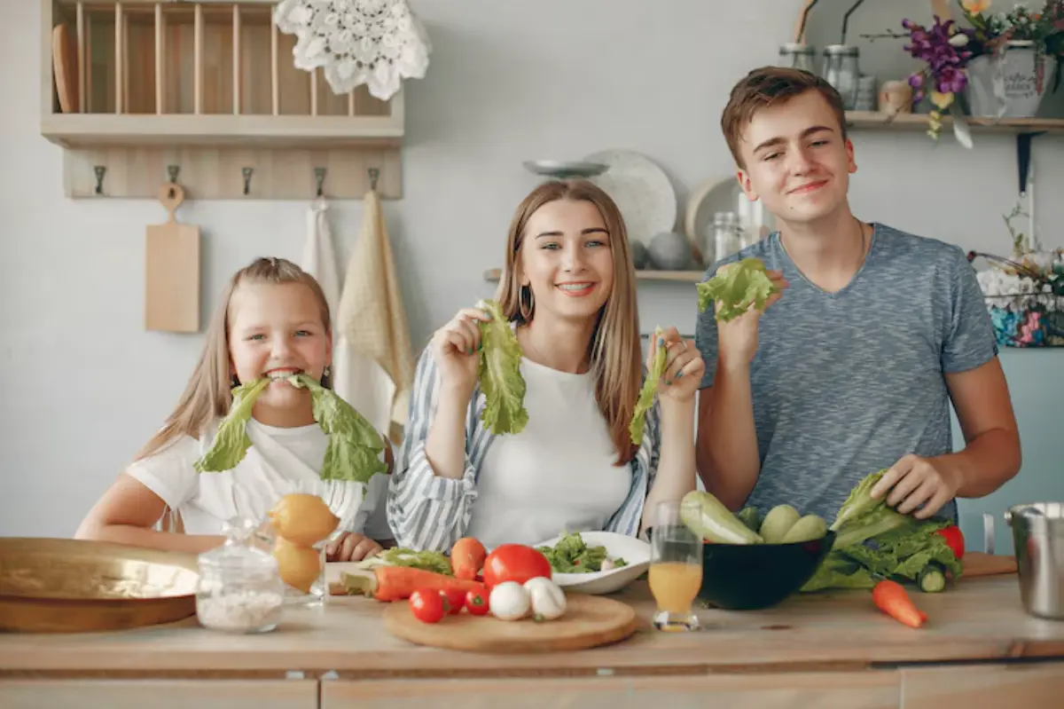 A family joyfully holds fresh lettuce in a bright kitchen, surrounded by colorful vegetables and a glass of juice on the table.