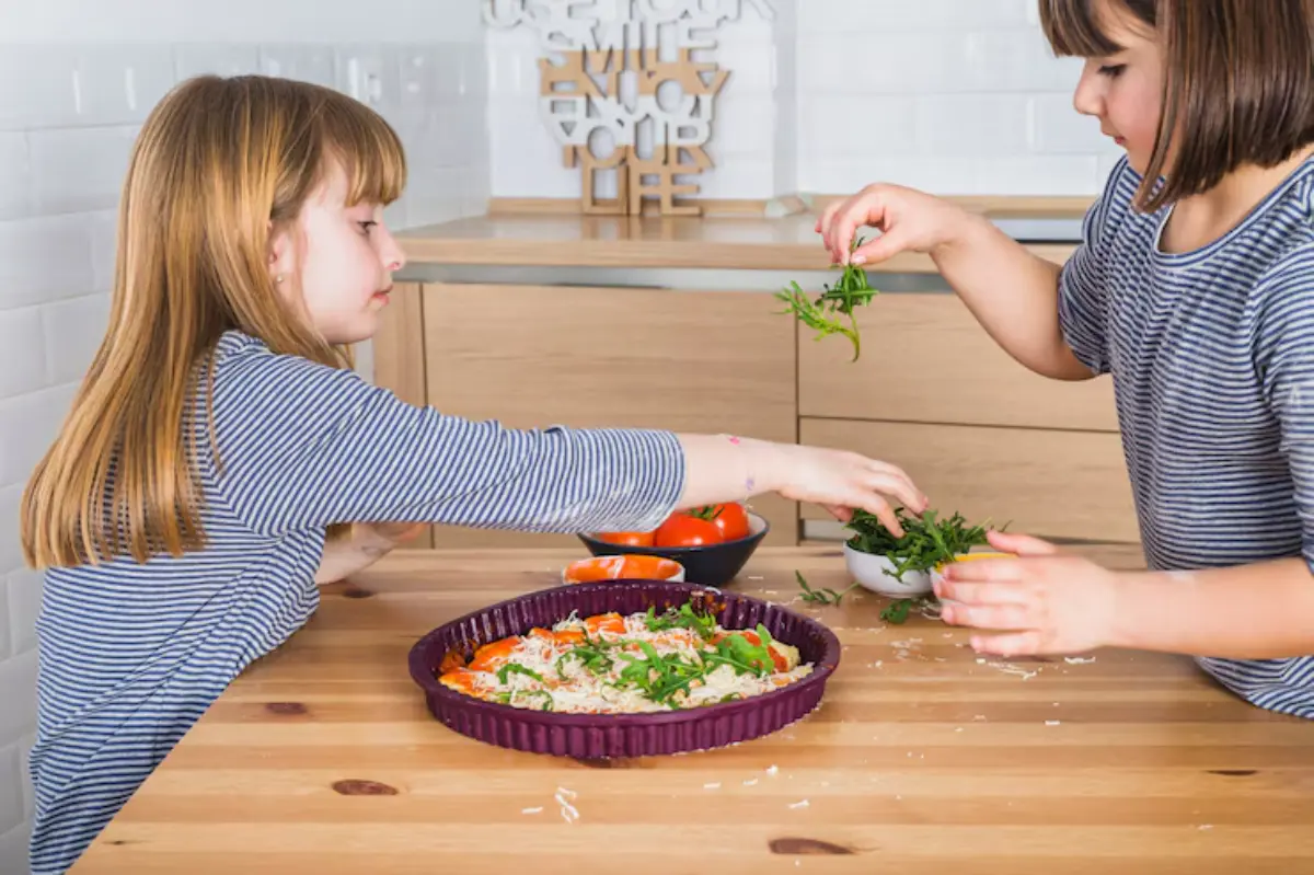 Two children are preparing a colorful dish in a modern kitchen, adding fresh ingredients to a purple baking dish on a wooden table.