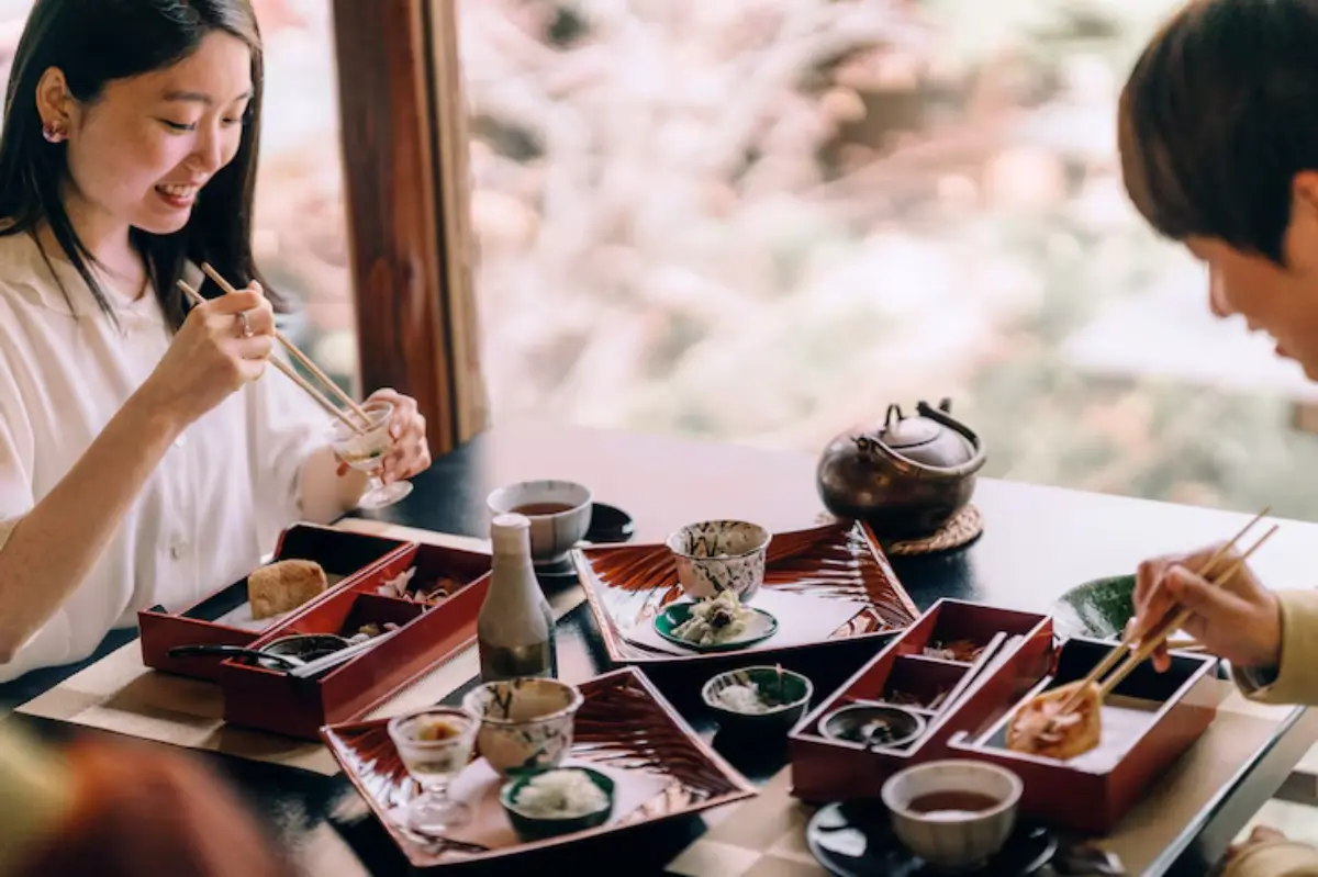 A table set with traditional Japanese dishes, including bento boxes and tea, as diners enjoy their meal with chopsticks.