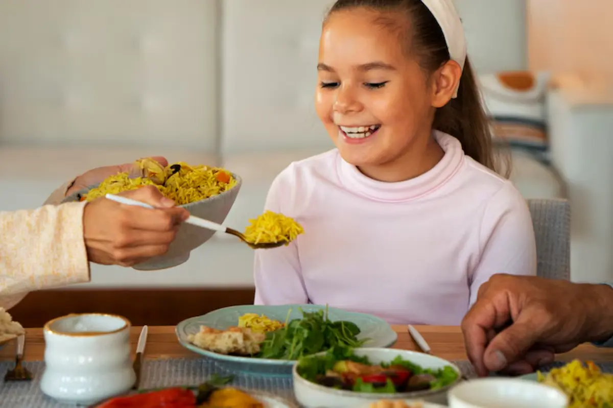 A hand serves a bowl of yellow rice to a child at a dining table with various dishes, creating a warm family meal setting.