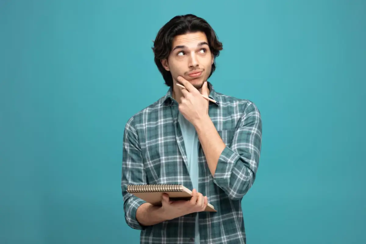 A person in a plaid shirt holds a notebook and a pen, deep in thought against a bright turquoise background.