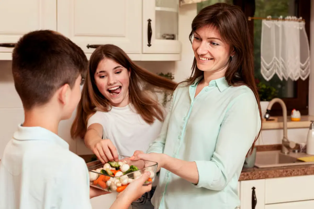 A woman in a light blue shirt hands a bowl of mixed vegetables to a boy while a girl reaches in to grab some.