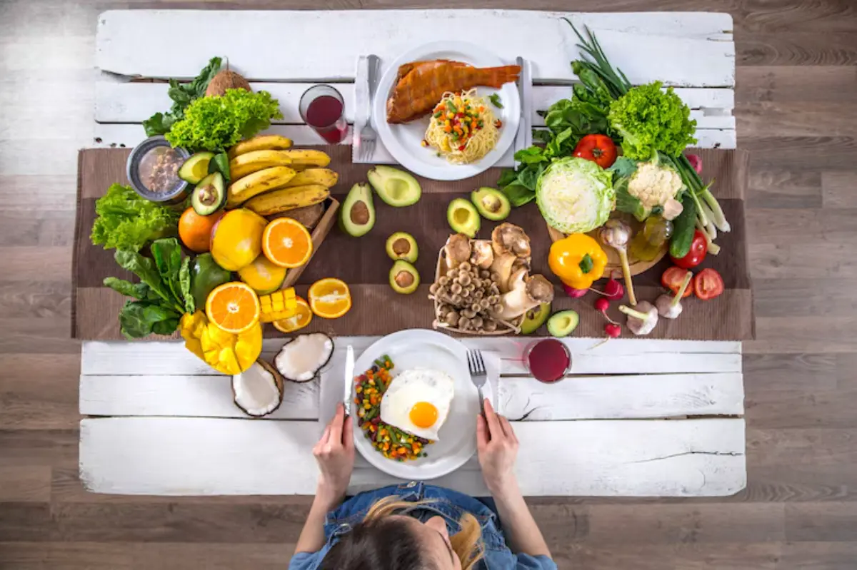 A vibrant table spread with a variety of fresh fruits, vegetables, and a plate of rice topped with a fried egg, enjoyed by a woman.