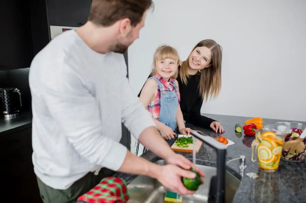 A man and a young girl are preparing vegetables at a modern kitchen counter, with fresh ingredients displayed around them.