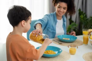 A young child pours cereal into a bowl at a bright breakfast table, surrounded by colorful plates, juice, and toast.