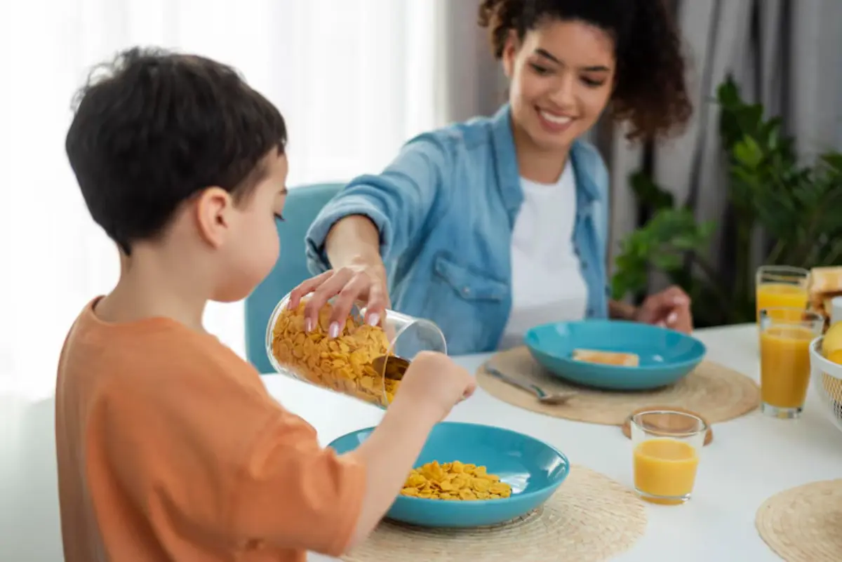A young child pours cereal into a bowl at a bright breakfast table, surrounded by colorful plates, juice, and toast.