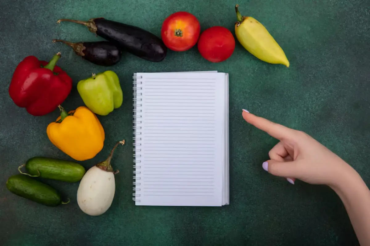 A blank notebook surrounded by colorful vegetables and a finger pointing, suggesting meal planning or cooking ideas.