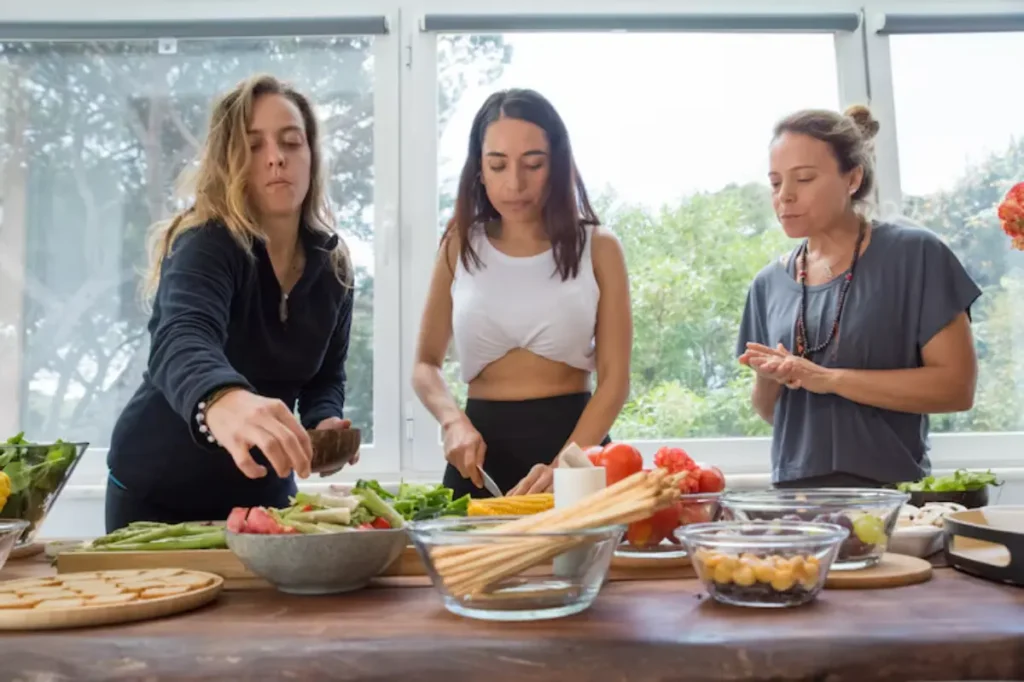 Three women are preparing meals in a bright kitchen. Fresh vegetables and ingredients are arranged on a wooden table.