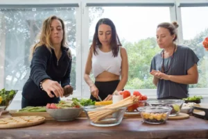 Three women are preparing meals in a bright kitchen. Fresh vegetables and ingredients are arranged on a wooden table.