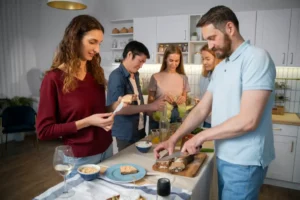A group of friends gather in a bright kitchen, enjoying food and drinks, while one person slices bread on a wooden board.