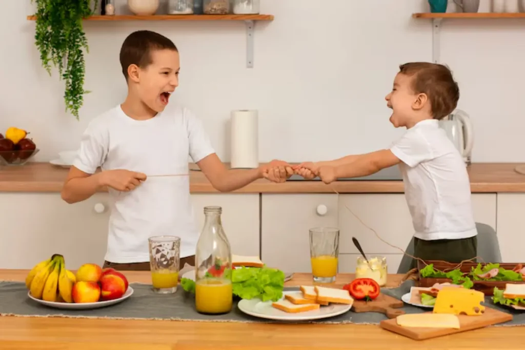 Two children play tug-of-war with a string in a bright kitchen, surrounded by fresh fruits, sandwiches, and drinks on the table.