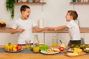 Two children play tug-of-war with a string in a bright kitchen, surrounded by fresh fruits, sandwiches, and drinks on the table.