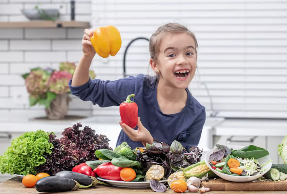 A child holds a yellow bell pepper in one hand and a red bell pepper in the other, surrounded by various fresh vegetables.