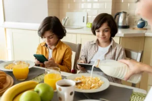 Two children sit at a breakfast table, one with a phone, the other with a tablet, surrounded by food like pasta, juice, and fruit.