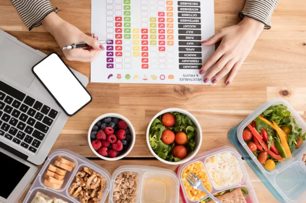 A person organizes meal plans with healthy food containers, a laptop, and a chart for tracking vegetables and fruits intake.