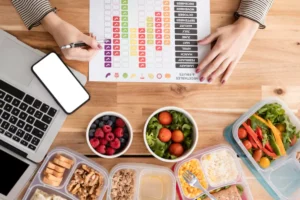 A person organizes meal plans with healthy food containers, a laptop, and a chart for tracking vegetables and fruits intake.