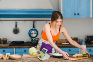 A woman in an orange top prepares food in a bright kitchen, with lettuce, chicken, and a tablet on the table.
