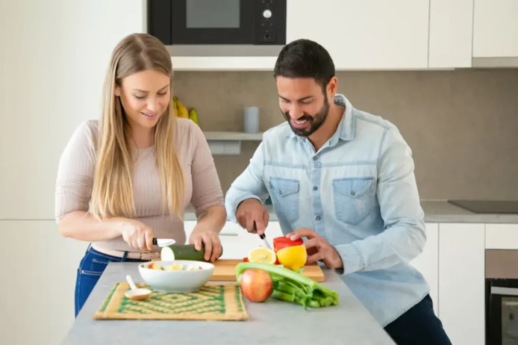 A woman and a man prepare food together in a modern kitchen, surrounded by fresh vegetables and fruits on a countertop.