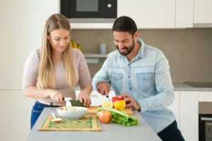 A woman and a man prepare food together in a modern kitchen, surrounded by fresh vegetables and fruits on a countertop.