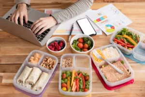 A person types on a laptop at a wooden table surrounded by healthy meal prep boxes, a phone, and colorful charts.