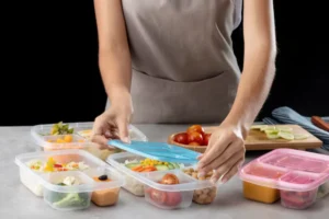 A person in an apron seals meal prep containers filled with colorful vegetables and fruits, preparing for storage on a kitchen counter.