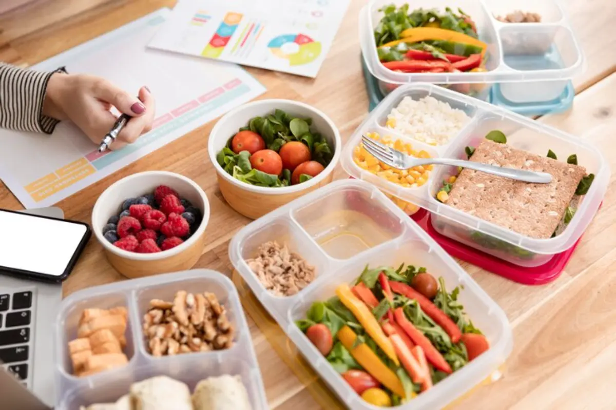 A workspace with healthy meal prep, featuring colorful salads, fruits, nuts, and a planning sheet with a hand writing notes.
