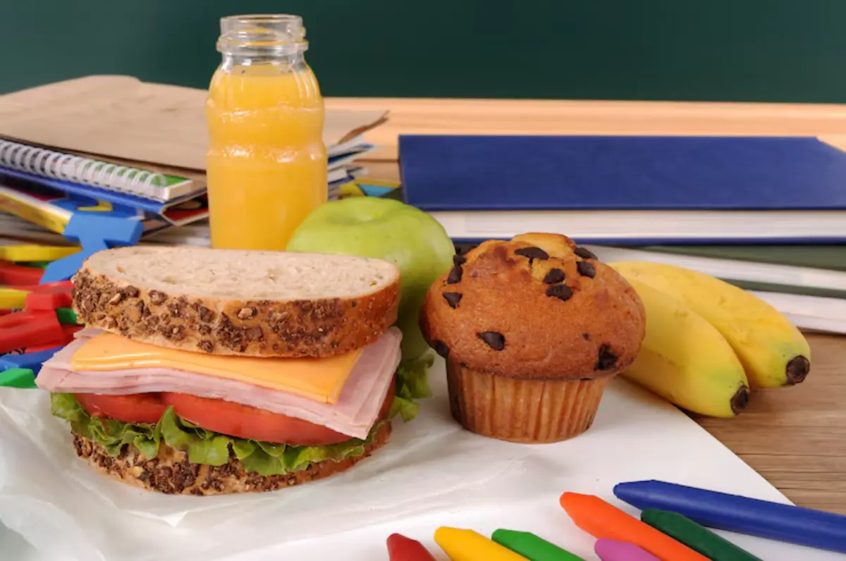 A packed lunch featuring a sandwich, muffin, fruit, and juice next to colorful crayons, on a desk surrounded by school supplies.