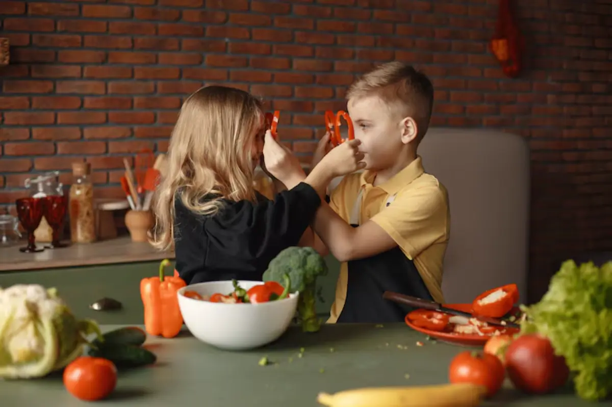 Two children playfully hold up sliced bell peppers in a cozy kitchen filled with fresh vegetables and a rustic brick wall.