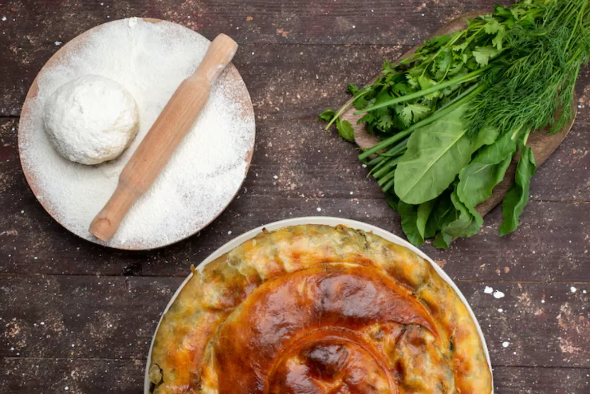 A golden-brown pie sits on a rustic table, surrounded by flour, a rolling pin, and fresh greens including dill and spinach.