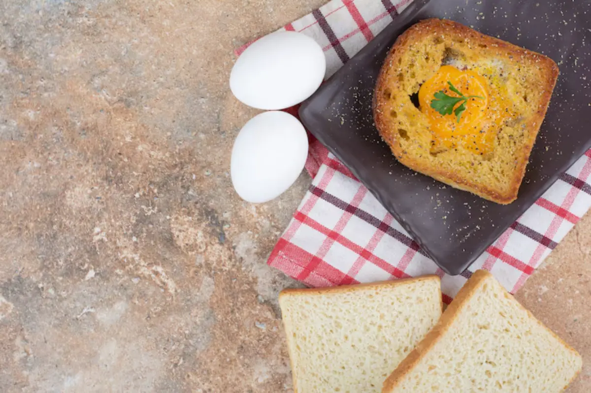 A slice of toasted bread with a runny egg yolk, garnished with parsley, beside two white eggs and slices of bread on a checkered cloth.